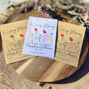 May include: Three seed packets with floral designs and the text "In Loving Memory." The packets are on a wooden tray. The text includes the name "Mary Rose Williams" and the years "1945-2025." The packets are labeled "Wildflower Seed Mix."