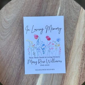 May include: A white card with the text "In Loving Memory" and "Plant These Seeds In Loving Memory". The card features watercolor illustrations of wildflowers and the name "Mary Rose Williams 1945-2025". The words "WILDFLOWER SEED MIX" are also printed on the card.