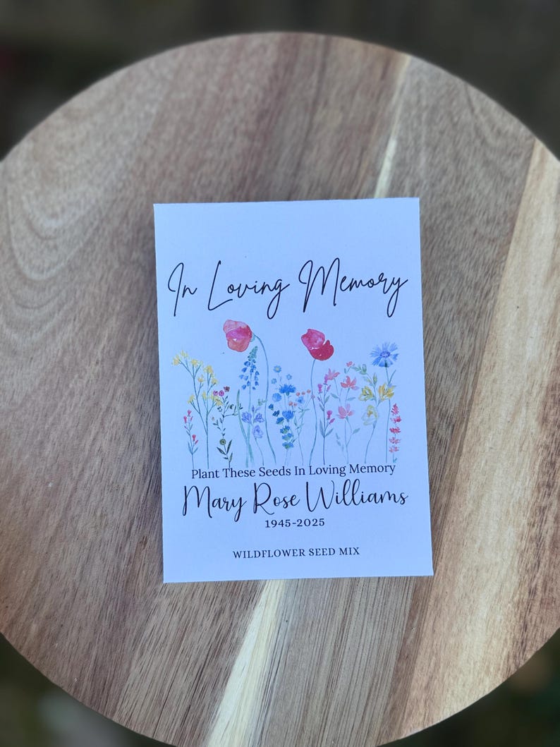 May include: A white card with the text "In Loving Memory" and "Plant These Seeds In Loving Memory". The card features watercolor illustrations of wildflowers and the name "Mary Rose Williams 1945-2025". The words "WILDFLOWER SEED MIX" are also printed on the card.
