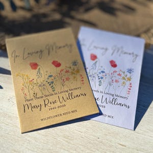 May include: Two seed packets with floral designs and the text "In Loving Memory." One packet is tan, the other white. Both packets read "Plant These Seeds In Loving Memory" and include a name and dates. The packets are labeled "WILDFLOWER SEED MIX."