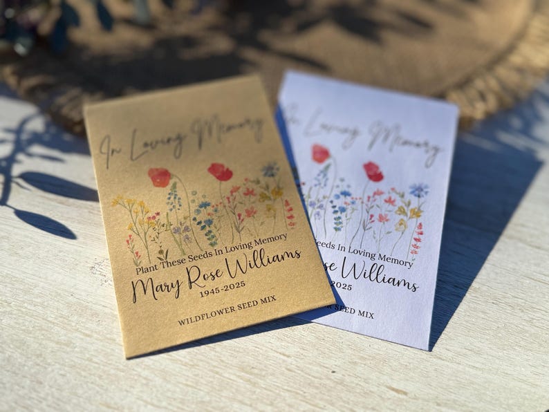 May include: Two seed packets with floral designs and the text "In Loving Memory." One packet is tan, the other white. Both packets read "Plant These Seeds In Loving Memory" and include a name and dates. The packets are labeled "WILDFLOWER SEED MIX."