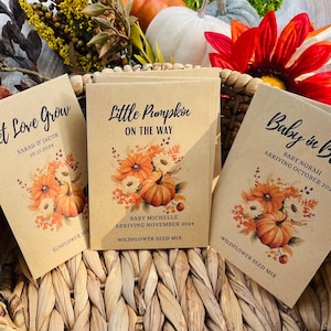 May include: Three brown paper seed packets with fall-themed designs. The packets have text that reads "Let Love Grow", "Little Pumpkin on the Way", and "Baby in Bloom". Each packet includes a floral design with pumpkins and the text "Wildflower Seed Mix" or "Sunflower Seed Mix".