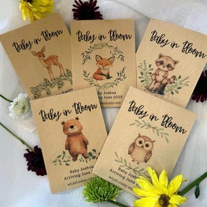 May include: Five brown paper seed packets with the text "Baby in Bloom" printed on them. Each packet features a different woodland animal illustration: a deer, a fox, a raccoon, a bear, and an owl. The packets also include the text "Baby Joshua Arriving June" and "Wildflower Seed Mix".