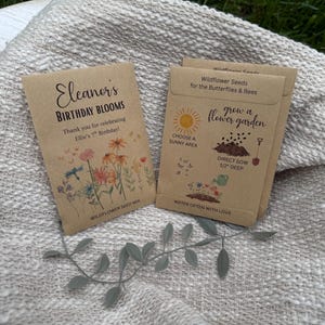 May include: Two seed packets on a textured, off-white blanket. One packet reads "Eleanor's Birthday Blooms" with floral illustrations. The other packet says "Wildflower Seeds for the Butterflies & Bees" with gardening instructions.