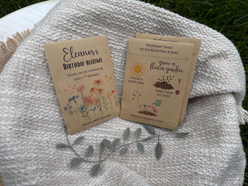 May include: Two seed packets on a textured, off-white blanket. One packet reads "Eleanor's Birthday Blooms" with floral illustrations. The other packet says "Wildflower Seeds for the Butterflies & Bees" with gardening instructions.