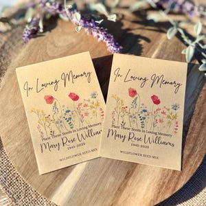 May include: Two kraft paper seed packets with the text "In Loving Memory" and "Mary Rose Williams 1945-2025". Each packet features a floral illustration with red poppies and other wildflowers. The packets are on a wooden tray.