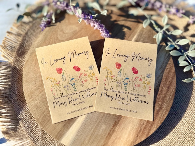 May include: Two kraft paper seed packets with the text "In Loving Memory" and "Mary Rose Williams 1945-2025". Each packet features a floral illustration with red poppies and other wildflowers. The packets are on a wooden tray.