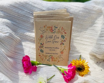 May include: A stack of kraft paper seed packets with floral designs and the text "Watch Our Love Grow" and names. The packets are surrounded by pink carnations, a white chrysanthemum, and a yellow sunflower, all on a white woven blanket.