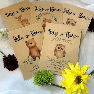 May include: Five brown paper envelopes with the text "Baby in Bloom" printed on them. Each envelope features a different woodland animal illustration: a deer, a fox, a raccoon, a bear, and an owl. The envelopes also include the text "Baby J Arriving" and "Baby Joshua Arriving June 2023" with the words "Wildflower Seed" printed below.