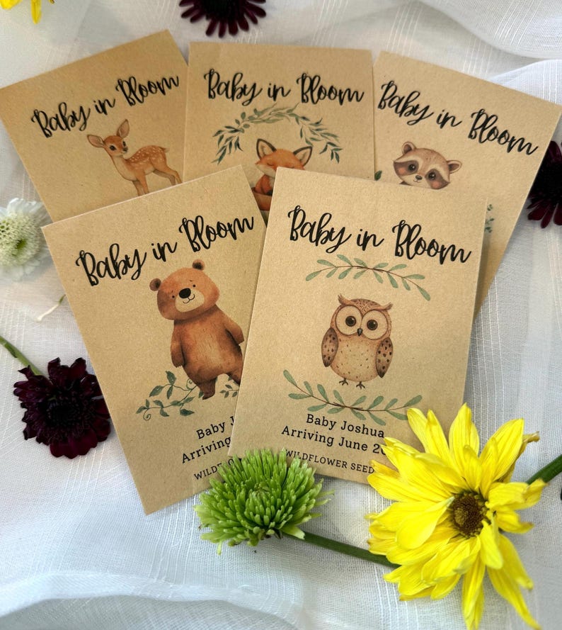 May include: Five brown paper envelopes with the text "Baby in Bloom" printed on them. Each envelope features a different woodland animal illustration: a deer, a fox, a raccoon, a bear, and an owl. The envelopes also include the text "Baby J Arriving" and "Baby Joshua Arriving June 2023" with the words "Wildflower Seed" printed below.
