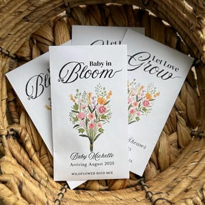 May include: A white seed packet with a floral design and the text "Baby in Bloom" and "Let Love Grow". The packet also includes the text "Baby Michelle Arriving August 2025 Wildflower Seed Mix".