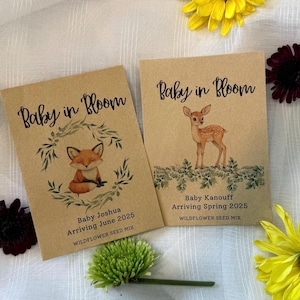 May include: Two brown paper packets with wildflower seed mix. The packets have a woodland theme with a fox and a deer on them. The text on the packets reads "Baby in Bloom" and includes the baby's name, arrival date, and "Wildflower Seed Mix".
