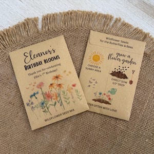 May include: Two brown paper seed packets with floral illustrations and text. One reads "Eleanor's Birthday Blooms" and "Wildflower Seed Mix." The other packet says "Wildflower Seeds for the Butterflies & Bees" and "grow a flower garden."