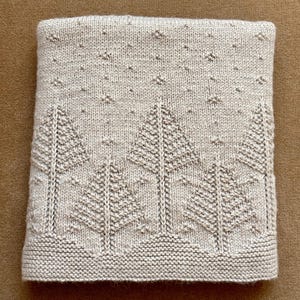 Könnte beinhalten: Eine beige gestrickte Babydecke mit einem Muster aus Bäumen und Sternen. Die Decke ist aus weichem, kuscheligem Garn gefertigt und perfekt zum Kuscheln.