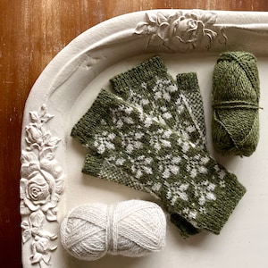 Pode incluir: Um par de luvas sem dedos de malha verde e branca com um padrão floral, juntamente com duas bolas de fio, uma verde e uma branca.