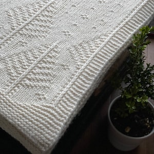 May include: A white knitted blanket with a textured pattern. The blanket is folded and has a white border.