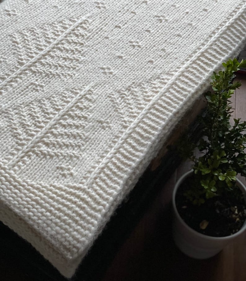 May include: A white knitted blanket with a textured pattern. The blanket is folded and has a white border.