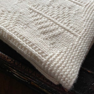 May include: A white knitted throw blanket with a textured pattern. The blanket is folded and lying on a dark surface.