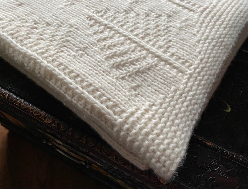 May include: A white knitted throw blanket with a textured pattern. The blanket is folded and lying on a dark surface.