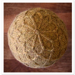 May include: A close-up of a knitted hat with a brown and gold color scheme. The hat has a unique pattern with a flower-like design in the center.