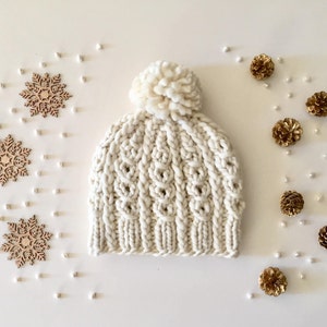 May include: A white knitted beanie with a pom pom on top. The beanie is surrounded by small wooden snowflakes and gold pine cones.