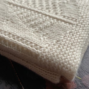 May include: A white knitted blanket with a textured pattern. The blanket is folded and the edge is visible.