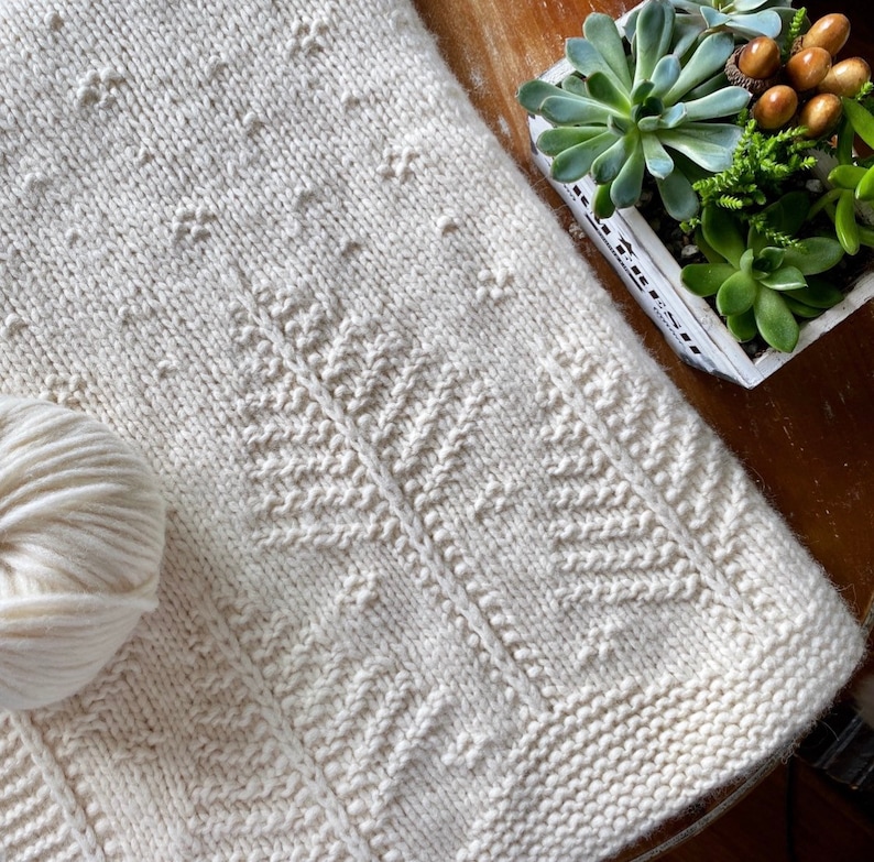 May include: A white knitted blanket with a repeating pattern of stylized trees. The blanket is made with a soft, textured yarn and has a subtle sheen.