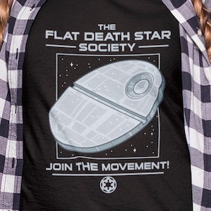Könnte beinhalten: Schwarzes T-Shirt mit dem Text "THE FLAT DEATH STAR SOCIETY" und einer Illustration eines flachen Todessterns. Das Shirt hat auch den Text "JOIN THE MOVEMENT!" und ein Symbol. Die Person trägt ein schwarz-weiß kariertes Hemd.