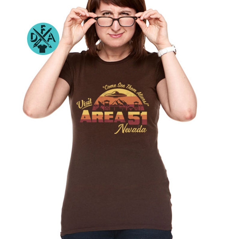 May include: A brown t-shirt with a retro-style graphic design featuring a sunset, a UFO, and the text "Visit Area 51 Nevada" with the tagline "Come See Them Aliens!"