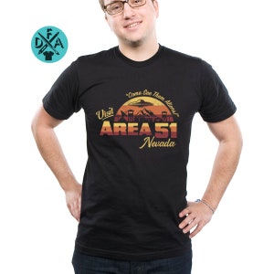 May include: Black t-shirt with a retro-style graphic of Area 51, Nevada. The graphic features a sunset, a UFO, and the text "Visit Area 51, Nevada" and "Come See Them Aliens".