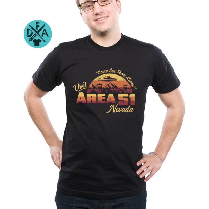 May include: Black t-shirt with a retro-style graphic of Area 51, Nevada. The graphic features a sunset, a UFO, and the text "Visit Area 51, Nevada" and "Come See Them Aliens".