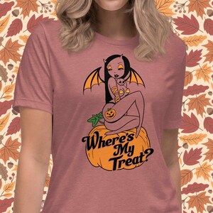 May include: Dusty rose t-shirt featuring a Halloween-themed graphic. The design includes a devilish woman with bat wings, sitting on a pumpkin, with the text "Where's My Treat?" in a stylized font. The background has a fall leaves pattern.