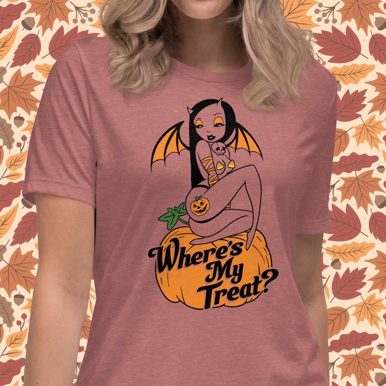 May include: Dusty rose t-shirt featuring a Halloween-themed graphic. The design includes a devilish woman with bat wings, sitting on a pumpkin, with the text "Where's My Treat?" in a stylized font. The background has a fall leaves pattern.