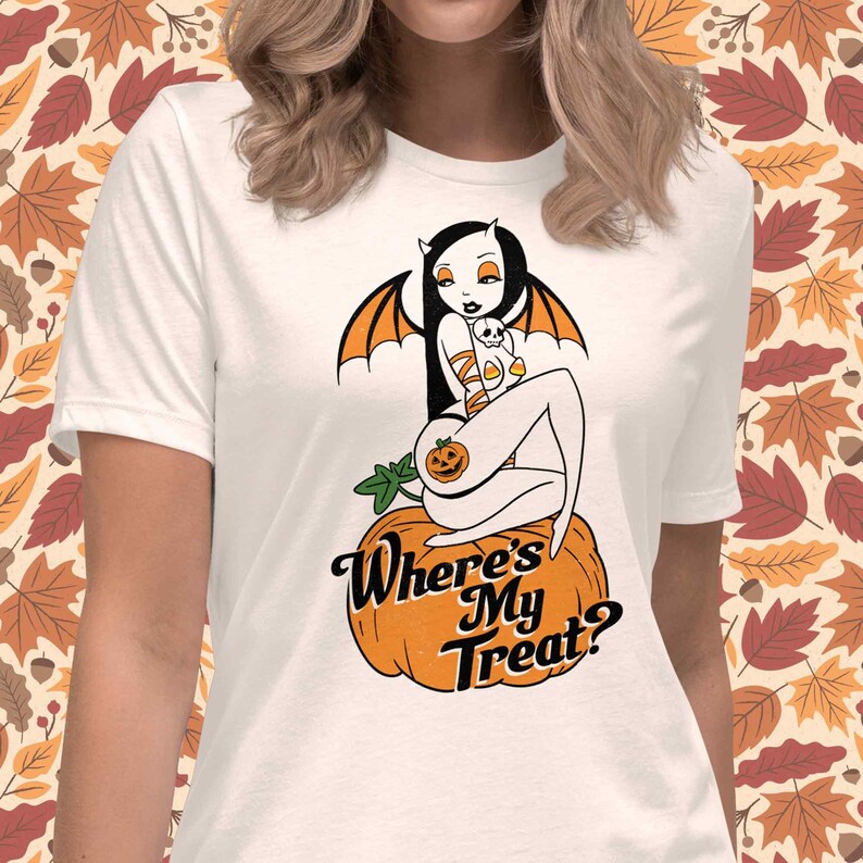 May include: White t-shirt featuring a Halloween-themed graphic. The design includes a seated figure with bat wings and horns, holding a skull and candy corn, atop an orange pumpkin. The text "Where's My Treat?" is below the pumpkin.