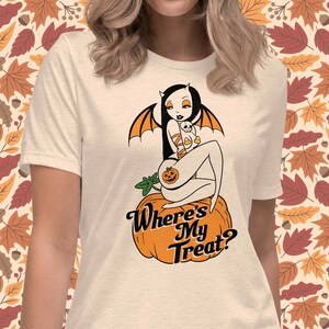 May include: Cream-colored t-shirt featuring a Halloween-themed graphic. The design includes a cartoon woman with bat wings and a pumpkin, sitting on a pumpkin. The text "Where's My Treat?" is below the graphic.