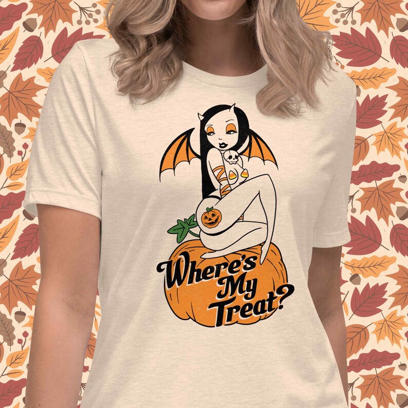 May include: Cream-colored t-shirt featuring a Halloween-themed graphic. The design includes a cartoon woman with bat wings and a pumpkin, sitting on a pumpkin. The text "Where's My Treat?" is below the graphic.