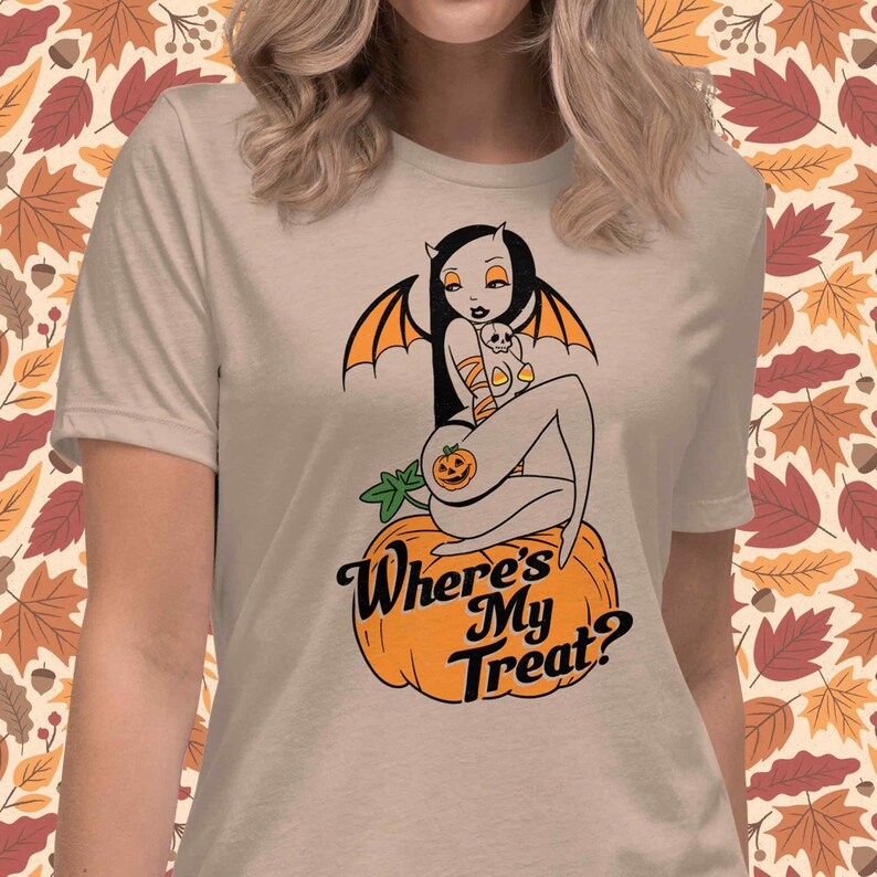 May include: Beige t-shirt featuring a Halloween-themed graphic. The design includes a seated devil-like figure with bat wings, a pumpkin, and the text "Where's My Treat?" in a stylized font. The background has a fall leaves pattern.