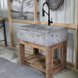 May include: A rectangular concrete sink basin with a mottled gray finish, mounted on a natural wood vanity base with four sturdy legs and a slatted lower shelf. A tall, black gooseneck faucet with a coiled spring neck is installed on the sink.