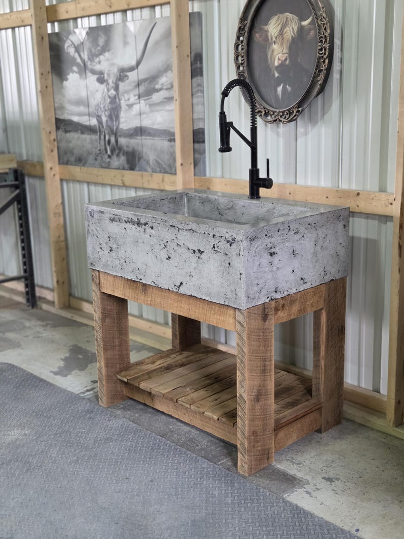 May include: A rectangular concrete sink basin with a mottled gray finish, mounted on a natural wood vanity base with four sturdy legs and a slatted lower shelf. A tall, black gooseneck faucet with a coiled spring neck is installed on the sink.