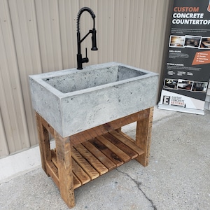 May include: A rectangular concrete sink basin with a textured gray surface and a matte black pull-down faucet. The sink is supported by a wooden base featuring four legs and a slatted lower shelf constructed from reclaimed-style brown timber planks.