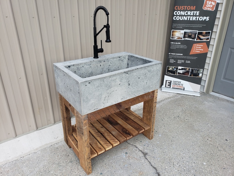 May include: A rectangular concrete sink basin with a textured gray surface and a matte black pull-down faucet. The sink is supported by a wooden base featuring four legs and a slatted lower shelf constructed from reclaimed-style brown timber planks.