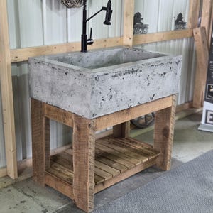 May include: A rectangular concrete sink basin with a textured gray surface, mounted on a wooden vanity base featuring four sturdy legs and a slatted lower shelf. A tall, black gooseneck faucet with a flexible pull-down sprayer is attached.