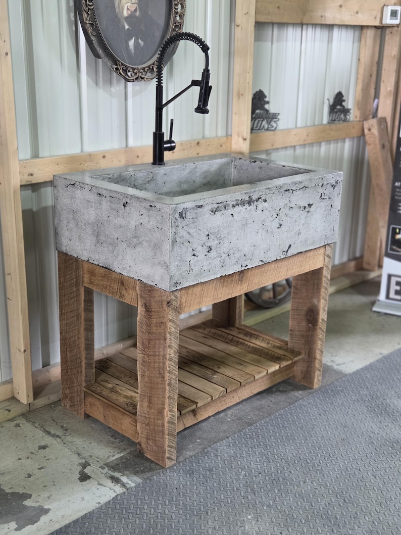 May include: A rectangular concrete sink basin with a textured gray surface, mounted on a wooden vanity base featuring four sturdy legs and a slatted lower shelf. A tall, black gooseneck faucet with a flexible pull-down sprayer is attached.