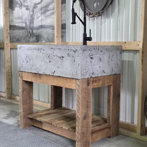 May include: A rectangular concrete sink basin with a mottled gray texture, mounted on a wooden vanity base with four sturdy legs and a slatted lower shelf. A tall, matte black gooseneck faucet with a pull-down sprayer is installed on the sink.