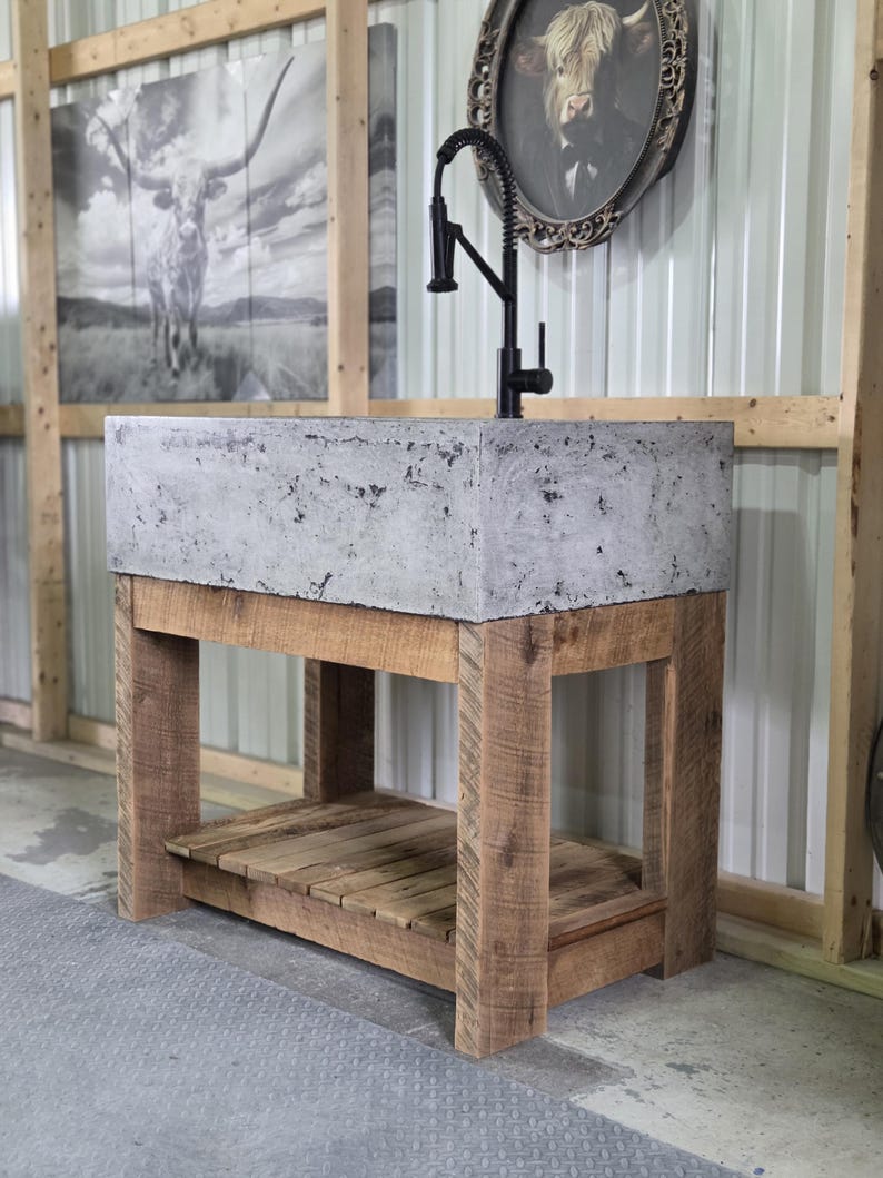 May include: A rectangular concrete sink basin with a mottled gray texture, mounted on a wooden vanity base with four sturdy legs and a slatted lower shelf. A tall, matte black gooseneck faucet with a pull-down sprayer is installed on the sink.