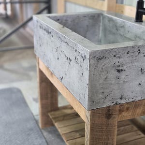 May include: A rectangular concrete sink with a mottled gray finish featuring a textured, porous surface. The basin is mounted on a base constructed from light-colored, unfinished wooden planks with a slatted lower shelf and a black metal faucet.