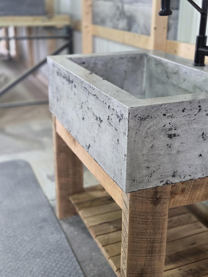 May include: A rectangular concrete sink with a mottled gray finish featuring a textured, porous surface. The basin is mounted on a base constructed from light-colored, unfinished wooden planks with a slatted lower shelf and a black metal faucet.