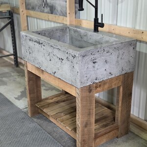 May include: A rectangular concrete sink with a mottled gray finish, mounted on a natural wood vanity base featuring four sturdy legs and a slatted lower shelf. A tall, black gooseneck faucet with a pull-down sprayer is installed on the sink deck.