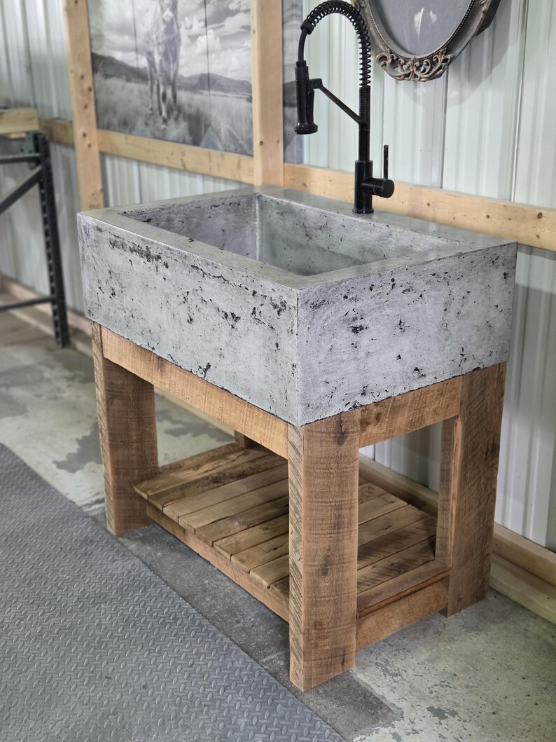 May include: A rectangular concrete sink with a mottled gray finish, mounted on a natural wood vanity base featuring four sturdy legs and a slatted lower shelf. A tall, black gooseneck faucet with a pull-down sprayer is installed on the sink deck.