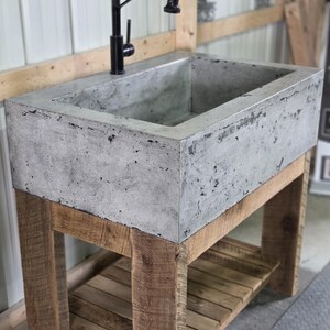 May include: A rectangular concrete farmhouse sink with a textured gray finish, mounted on a wooden vanity base with a slatted lower shelf. A tall, black gooseneck faucet with a pull-down sprayer is installed directly into the concrete basin.